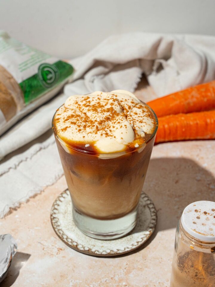 A glass tumbler with iced carrot cake latte topped with vanilla cream cheese foam and sprinkled with cinnamon. The glass is on a ceramic coaster on a tan counter with a white cloth napkin and carrots.