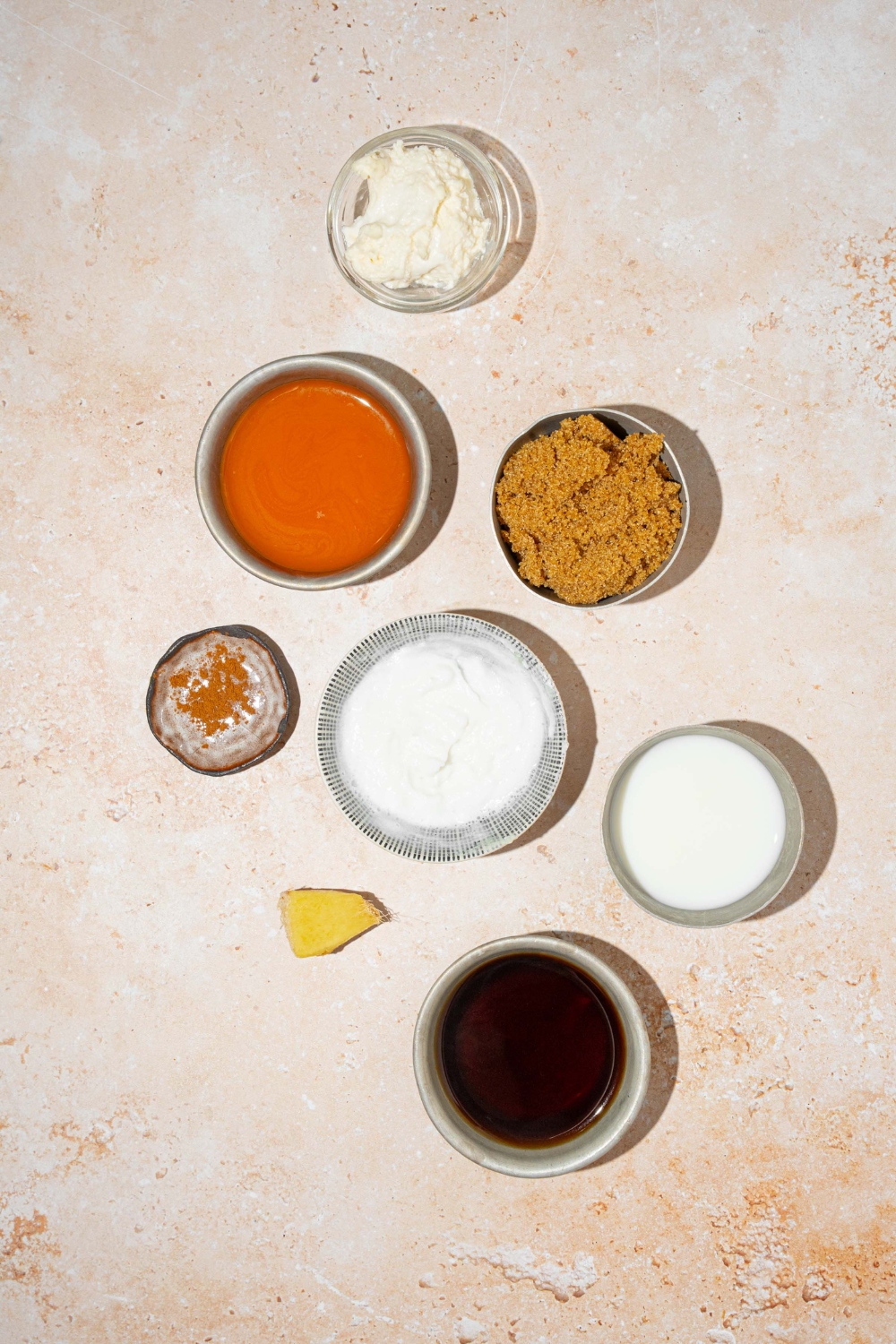 A tan counter with several bowls in various sizes containing ingredients to make carrot cake latte including carrot juice, cream cheese, brown sugar, heavy cream, skim milk, vanilla extract, and ginger.