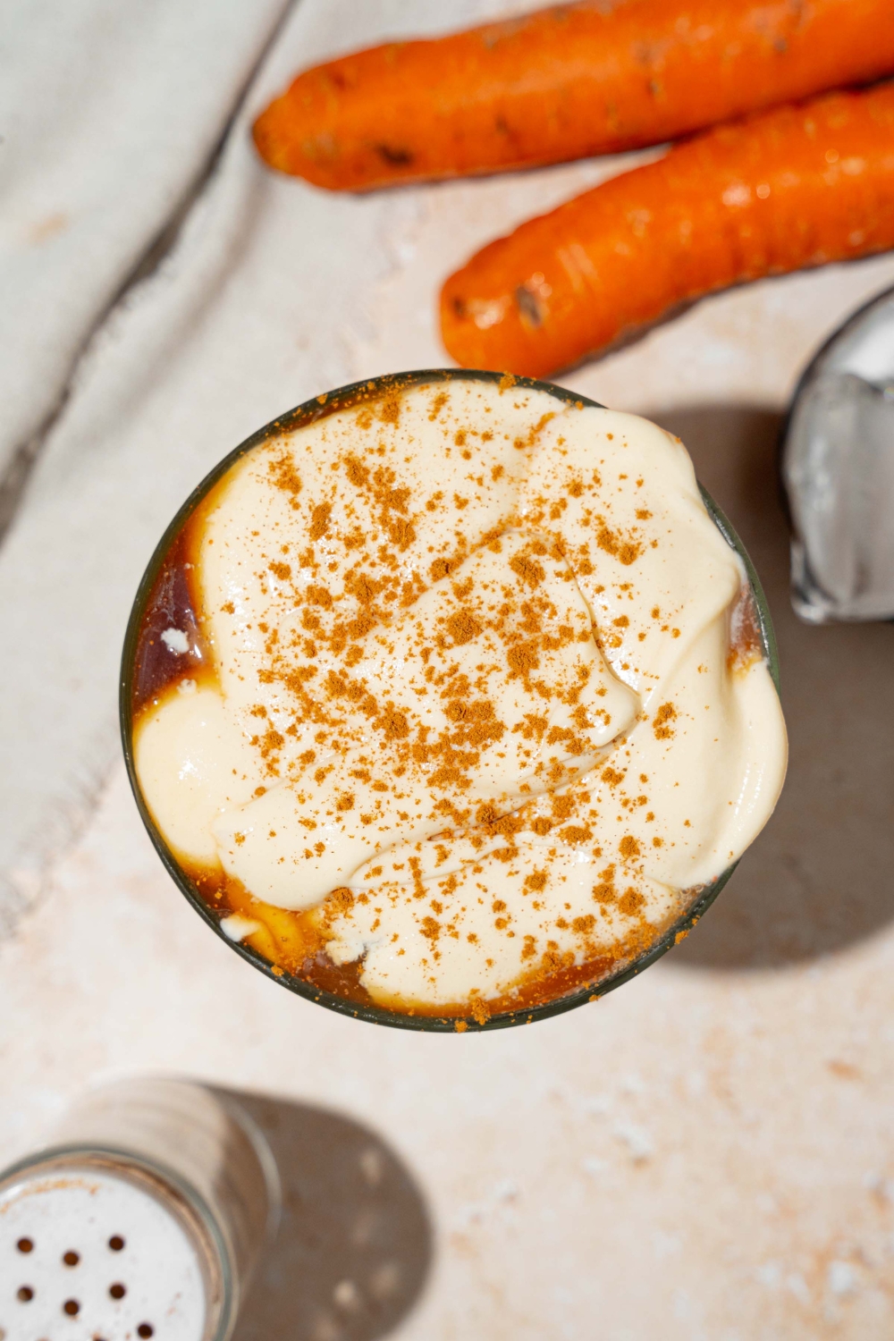 A glass tumbler with iced carrot cake latte topped with vanilla cream cheese foam and sprinkled with cinnamon. The glass is on a ceramic coaster on a tan counter with a white cloth napkin and carrots.