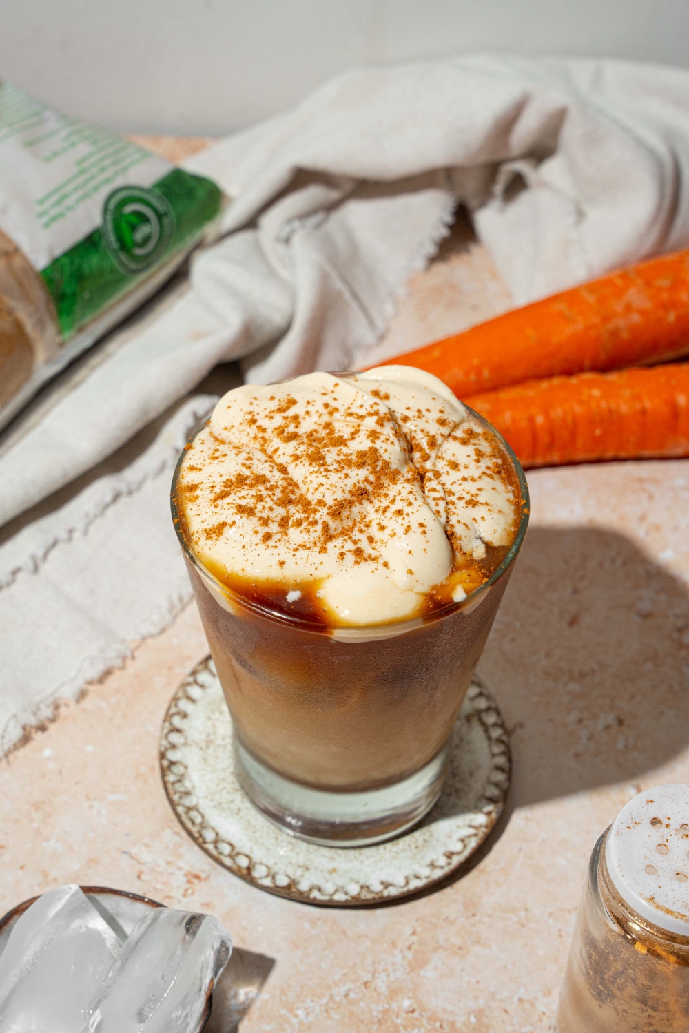 A glass tumbler with iced carrot cake latte topped with vanilla cream cheese foam and sprinkled with cinnamon. The glass is on a ceramic coaster on a tan counter with a white cloth napkin and carrots.