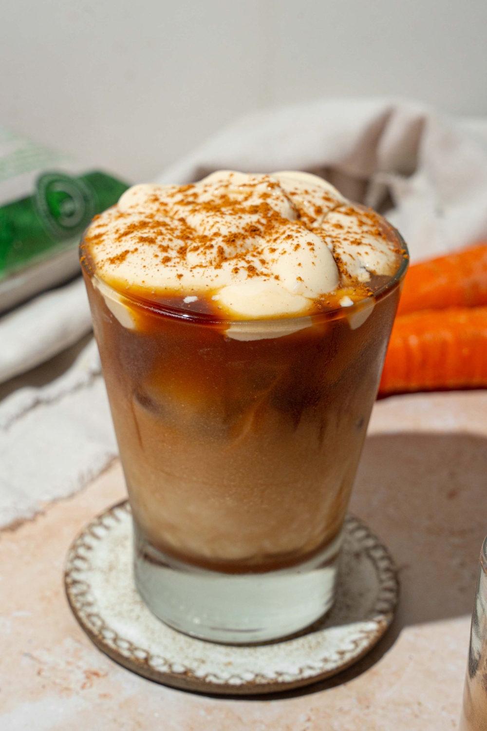 A glass tumbler with iced carrot cake latte topped with vanilla cream cheese foam and sprinkled with cinnamon. The glass is on a ceramic coaster on a tan counter with a white cloth napkin and carrots.