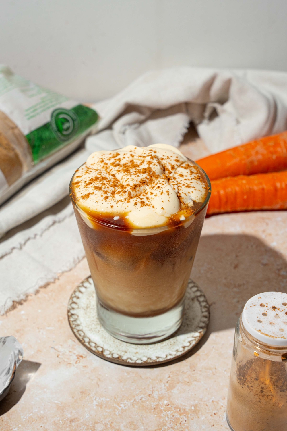 A glass tumbler with iced carrot cake latte topped with vanilla cream cheese foam and sprinkled with cinnamon. The glass is on a ceramic coaster on a tan counter with a white cloth napkin and carrots.