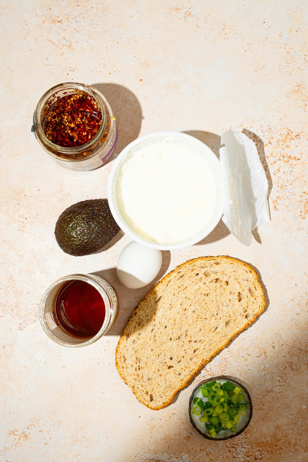 A tan counter with ingredients to make chili crisp avocado toast including a slice of bread, egg, cottage cheese, chili crisp oil, honey, and avocado.