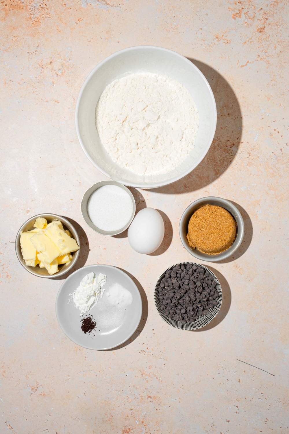 A tan counter with several bowls in various sizes containing ingredients to make chocolate chip cookie fries including flour, butter, brown sugar, sugar, egg, chocolate chips, and more.