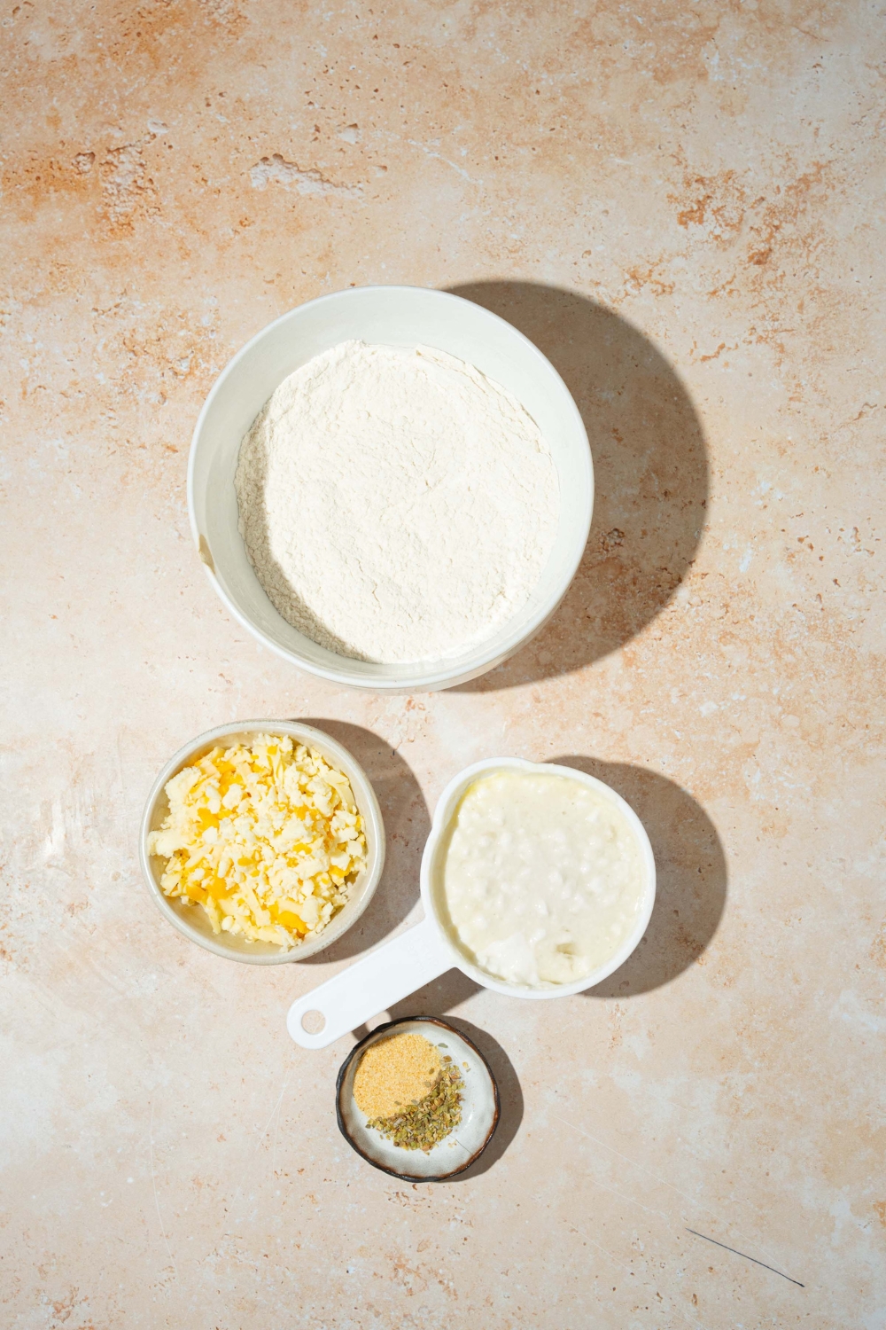 A tan counter with several bowls containing ingredients to make cottage cheese breadsticks including cottage cheese, shredded cheese, and flour.