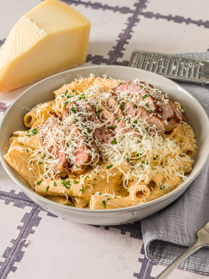 A white bowl with cajun steak pasta garnished with shredded cheese and fresh parsley. The bowl is on a tiled counter with a block of cheese and gray cloth napkin.