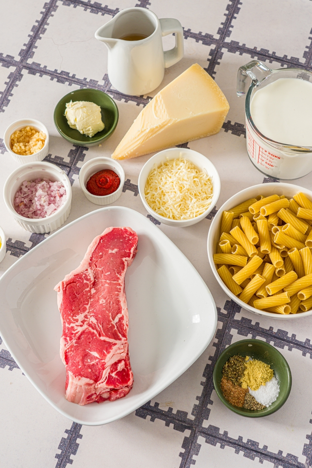 A tiled counter with several bowls in various sizes containing ingredients to make cajun steak pasta including steak, rigatoni pasta, heavy cream, shredded cheese, shallots, chicken broth, tomato paste, garlic, and seasonings.