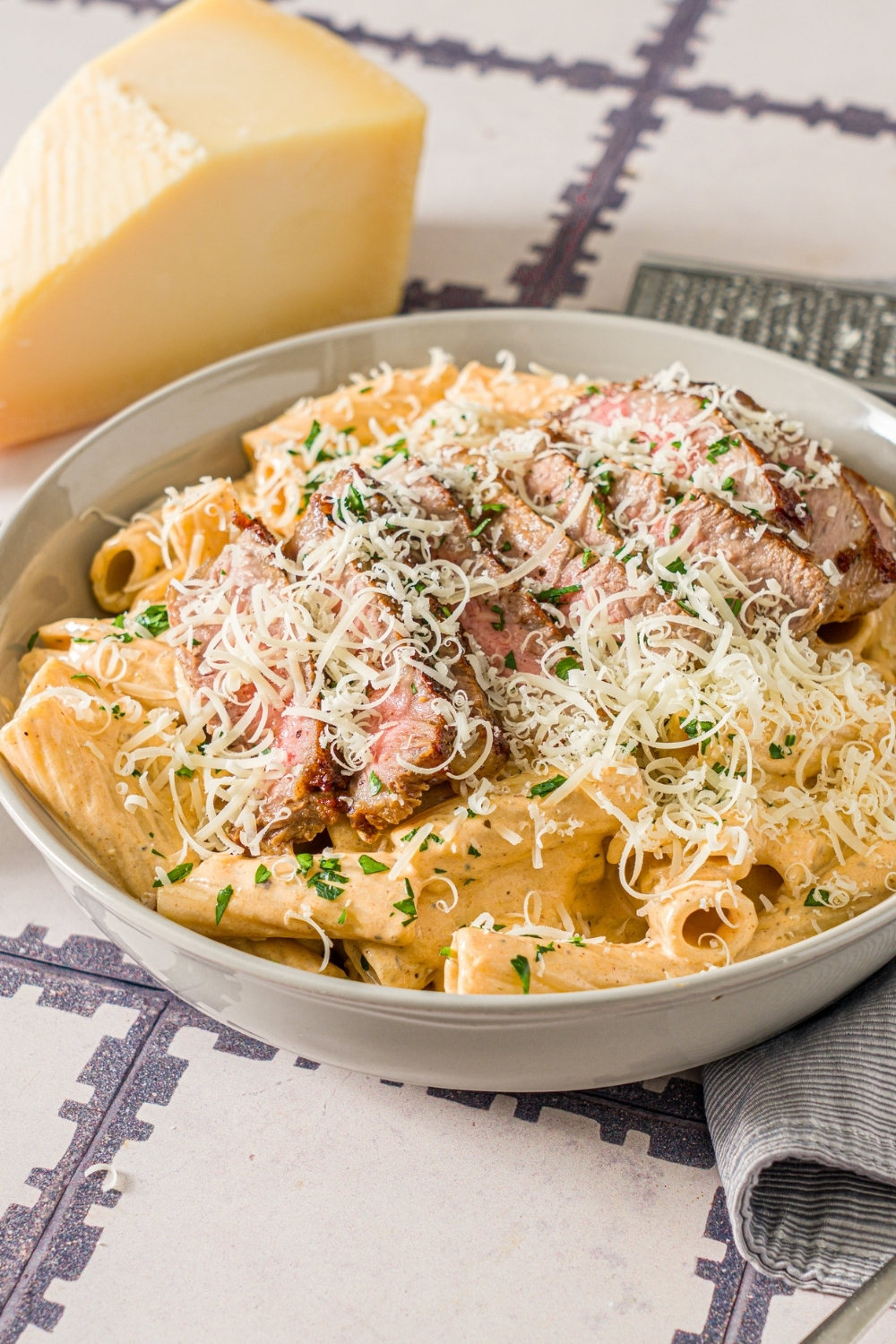 A white bowl with cajun steak pasta garnished with shredded cheese and fresh parsley. The bowl is on a tiled counter with a block of cheese and gray cloth napkin.