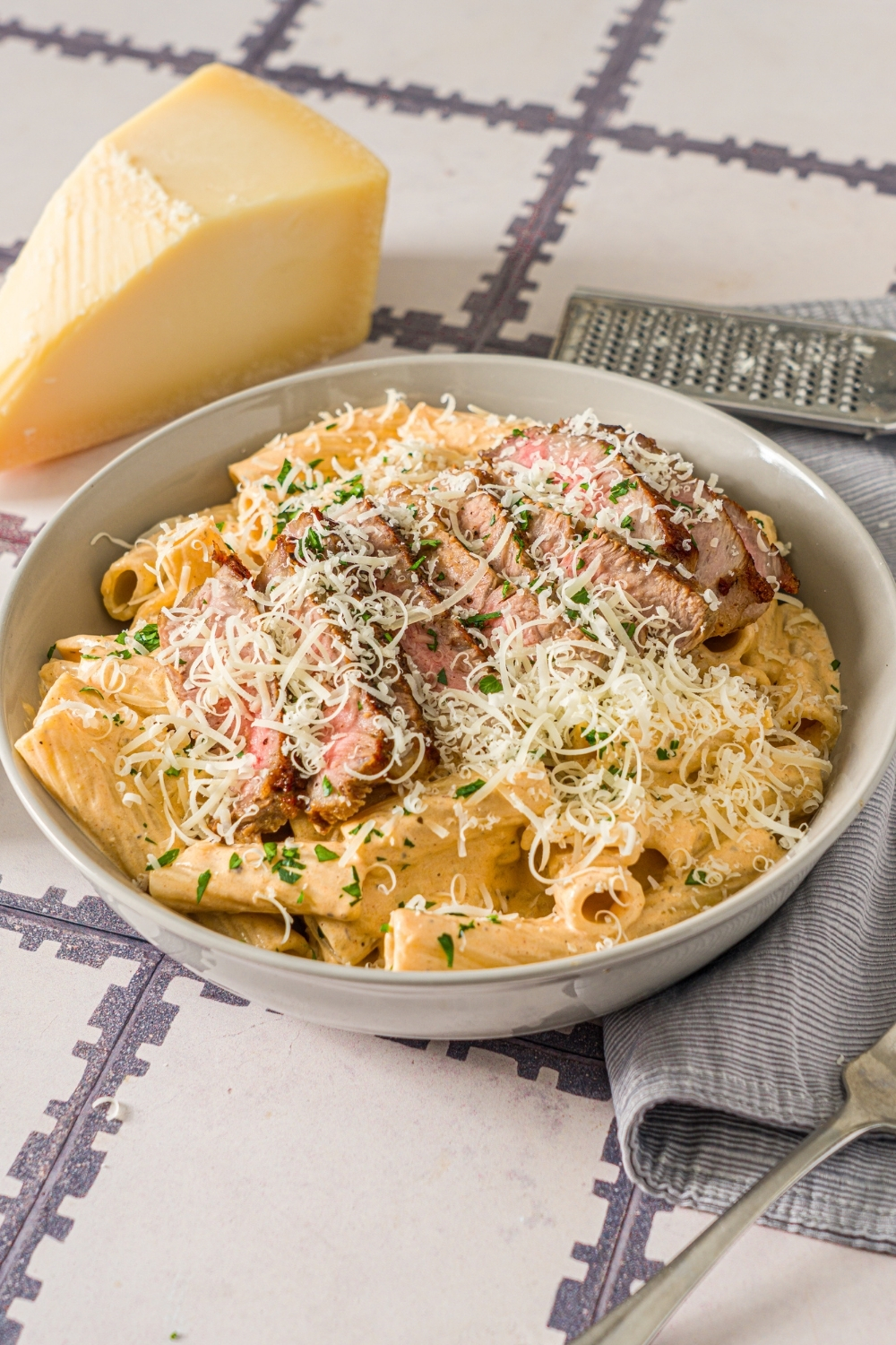 A white bowl with cajun steak pasta garnished with shredded cheese and fresh parsley. The bowl is on a tiled counter with a block of cheese and gray cloth napkin.