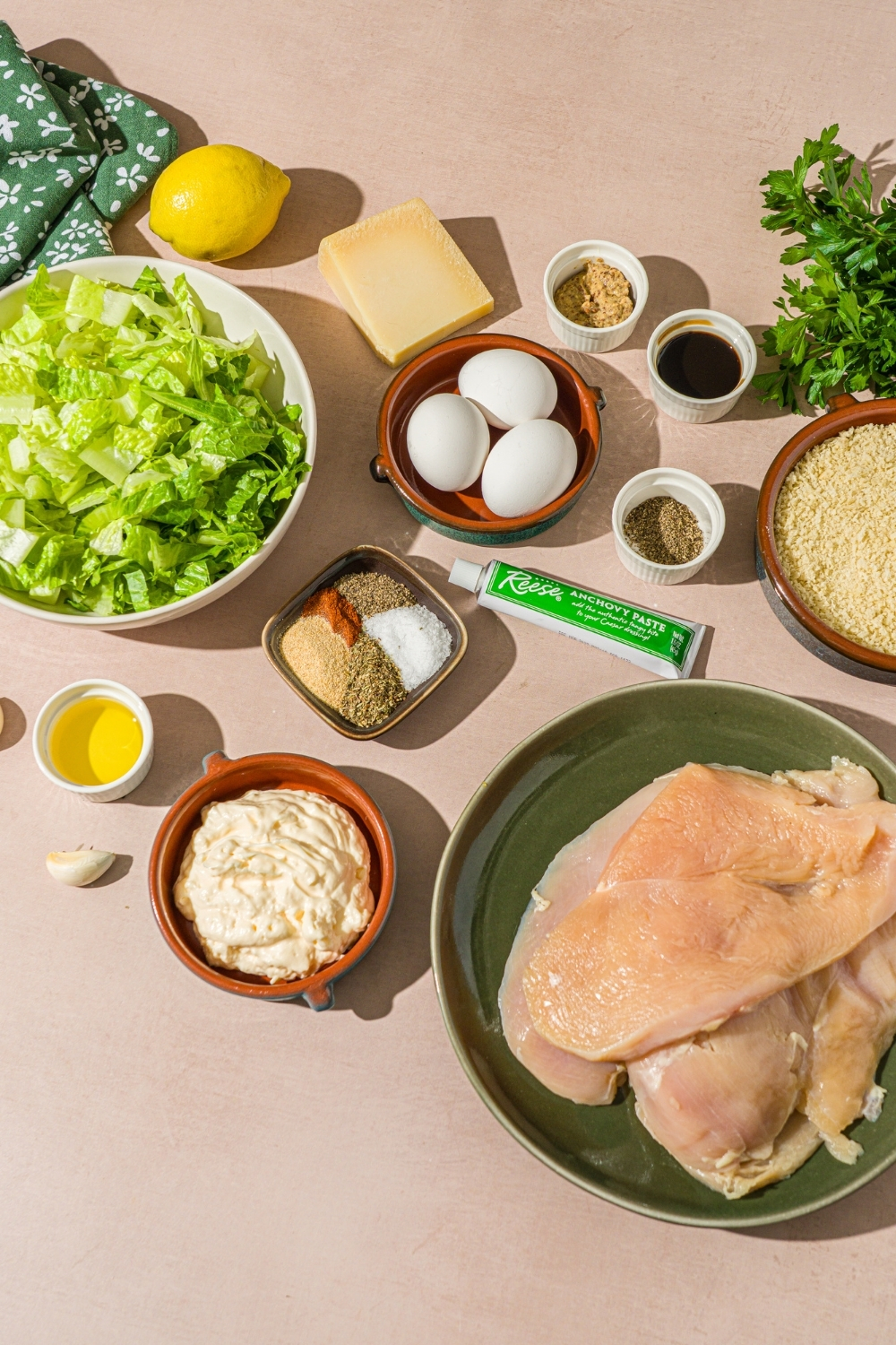 A tan counter with multiple bowls in various sizes containing ingredients to make crispy chicken caesar salad including chicken cutlets, eggs, romaine lettuce, shredded parmesan cheese, mayo, breadcrumbs, anchovy paste, and seasonings.