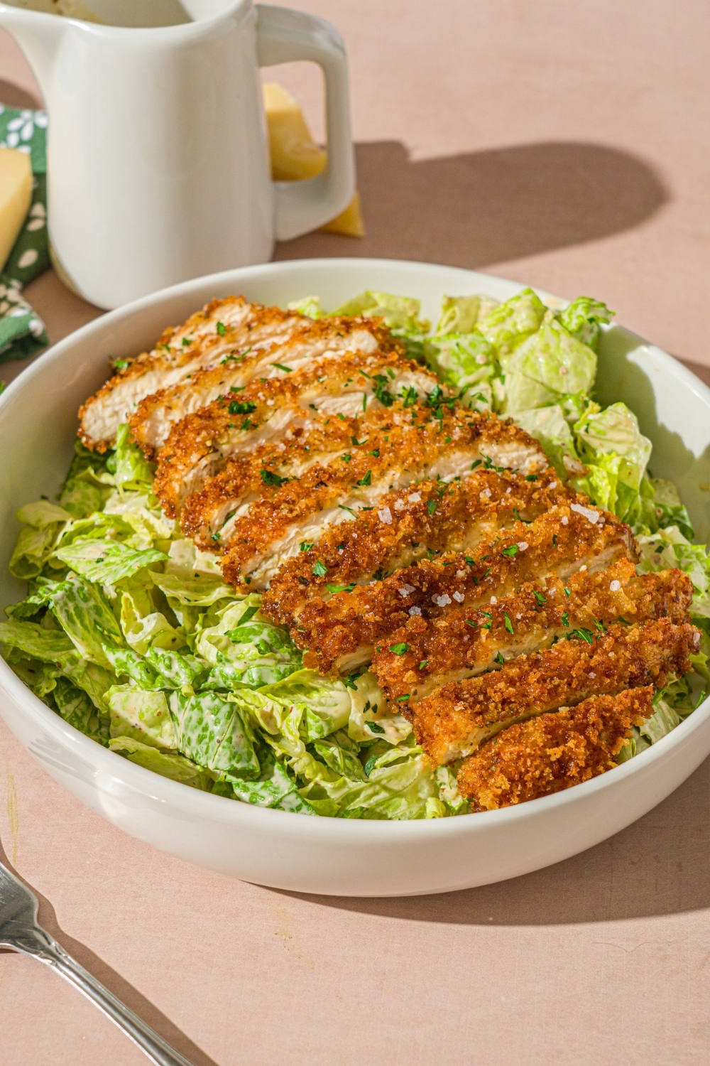 A white bowl with crispy chicken caesar salad garnished with fresh parsley. The bowl is on a tan counter with a creamer of dressing and wedge of parmesan cheese.
