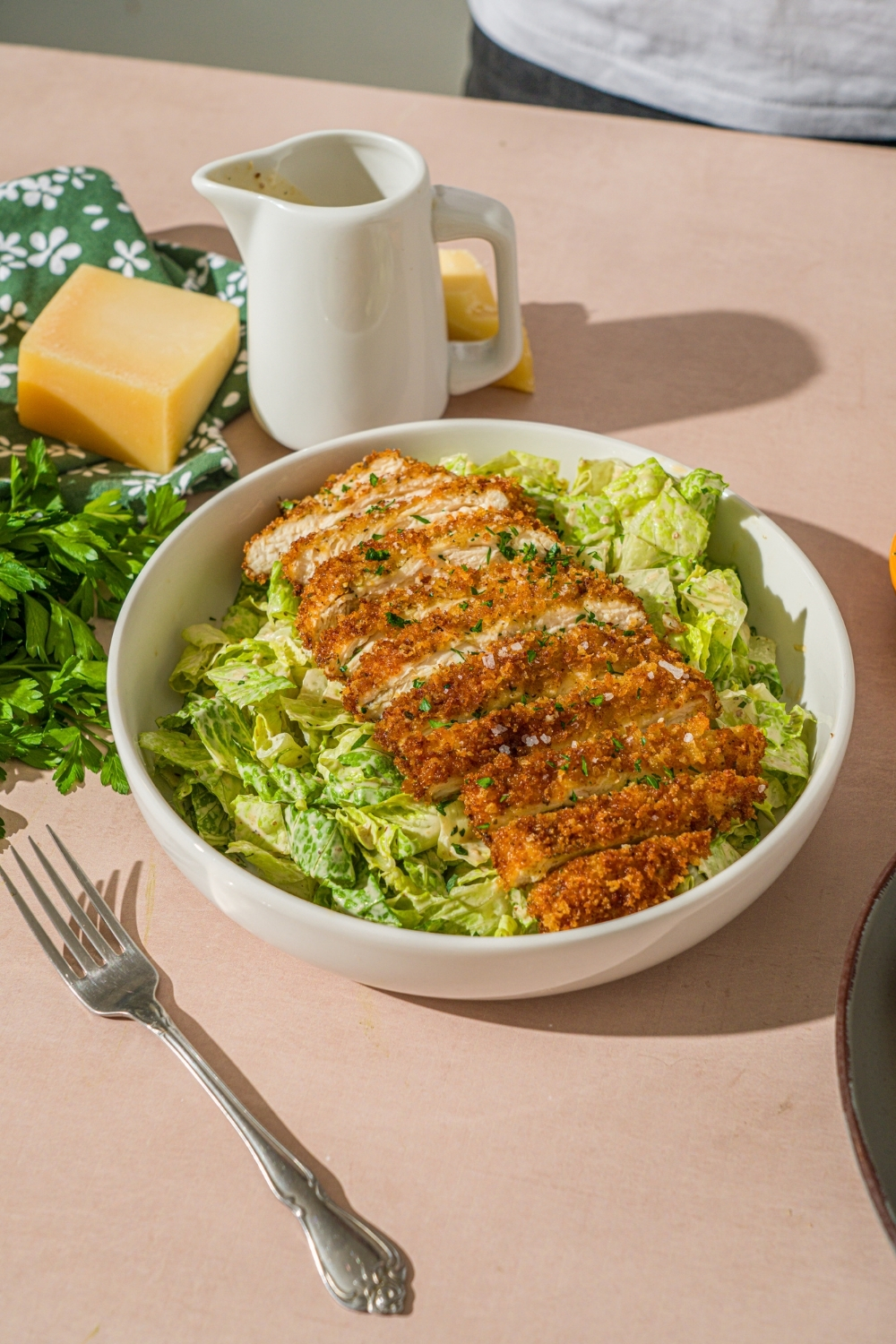 A white bowl with crispy chicken caesar salad garnished with fresh parsley. The bowl is on a tan counter with a creamer of dressing and wedge of parmesan cheese.