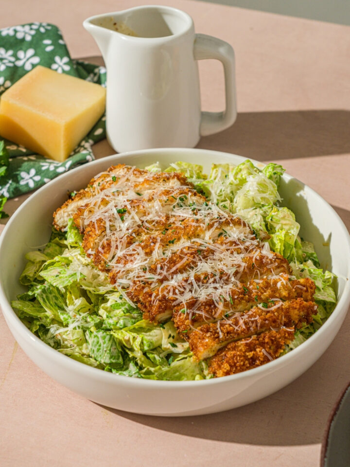 A white bowl with crispy chicken caesar salad garnished with fresh parsley and shredded parmesan. The bowl is on a tan counter with a creamer of dressing and wedge of parmesan cheese.