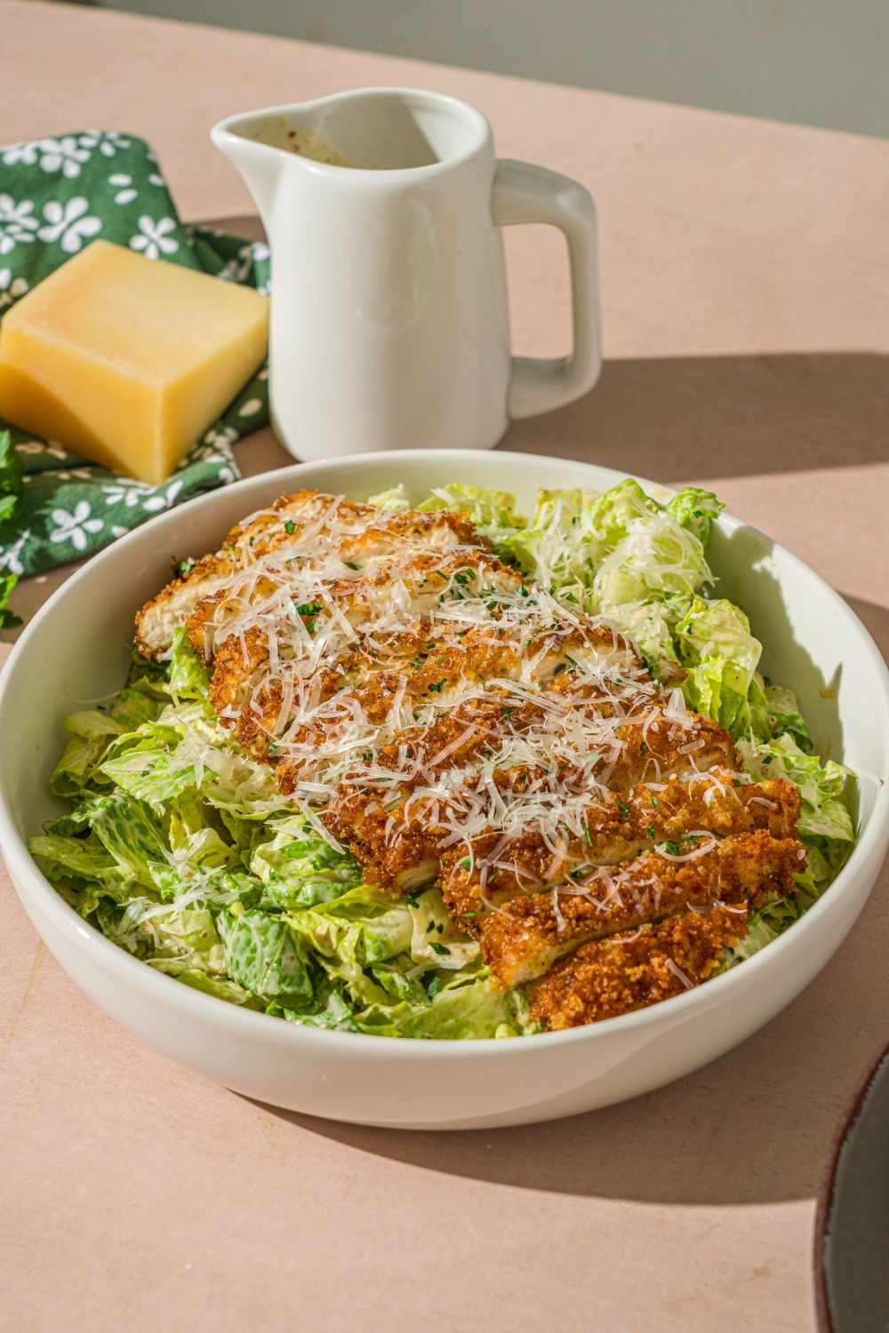 A white bowl with crispy chicken caesar salad garnished with fresh parsley and shredded parmesan. The bowl is on a tan counter with a creamer of dressing and wedge of parmesan cheese.