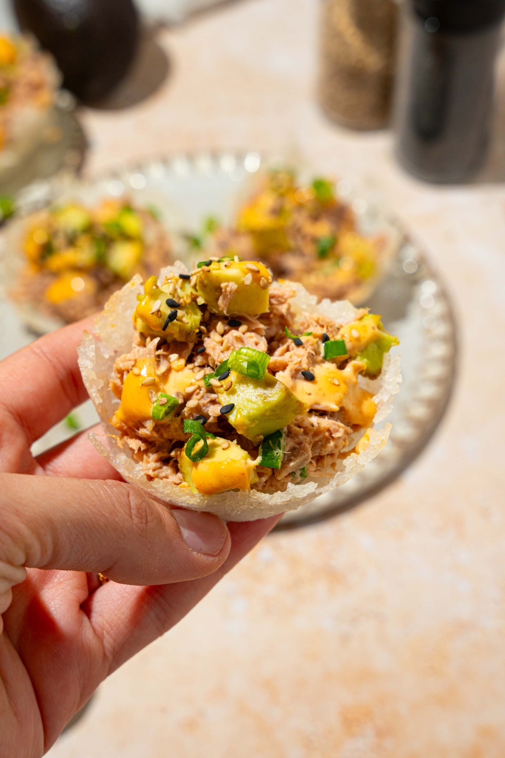 A close up of a hand holding a tuna rice up topped with avocado, sliced green onion, sesame seeds, and spicy mayo. There is a plate of tuna cups on a tan counter blurred in the background.