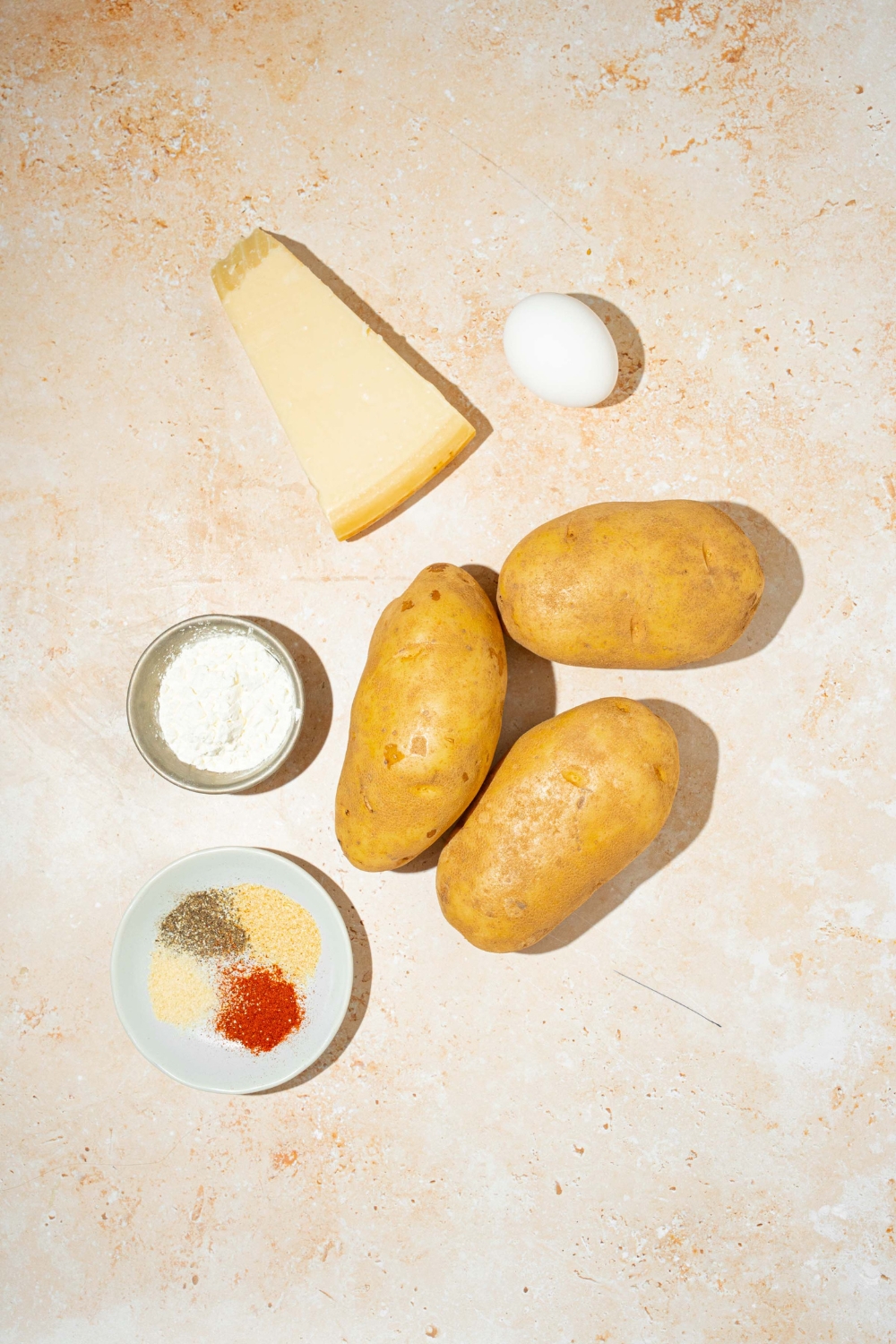 A tan counter with ingredients to make crispy parmesan hashbrowns including russet potatoes, parmesan cheese, egg, cornstarch, and seasonings.