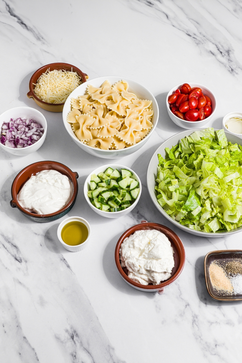 A marble counter with several bowls in various sizes containing ingredients to make crispy pasta salad including bowtie pasta, shredded lettuce, tomatoes, cucumber, yogurt, sour cream, red onion, olive oil, parmesan cheese, and seasonings.