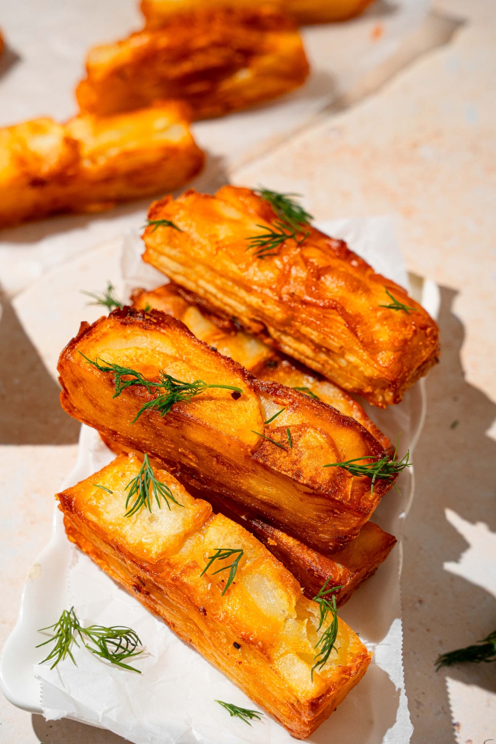 A white platter lined with parchment paper with several pieces of potato pave garnished with fresh thyme. The pieces are stacked on top of each other. The plate is on a tan counter with additional pieces of potato pave.