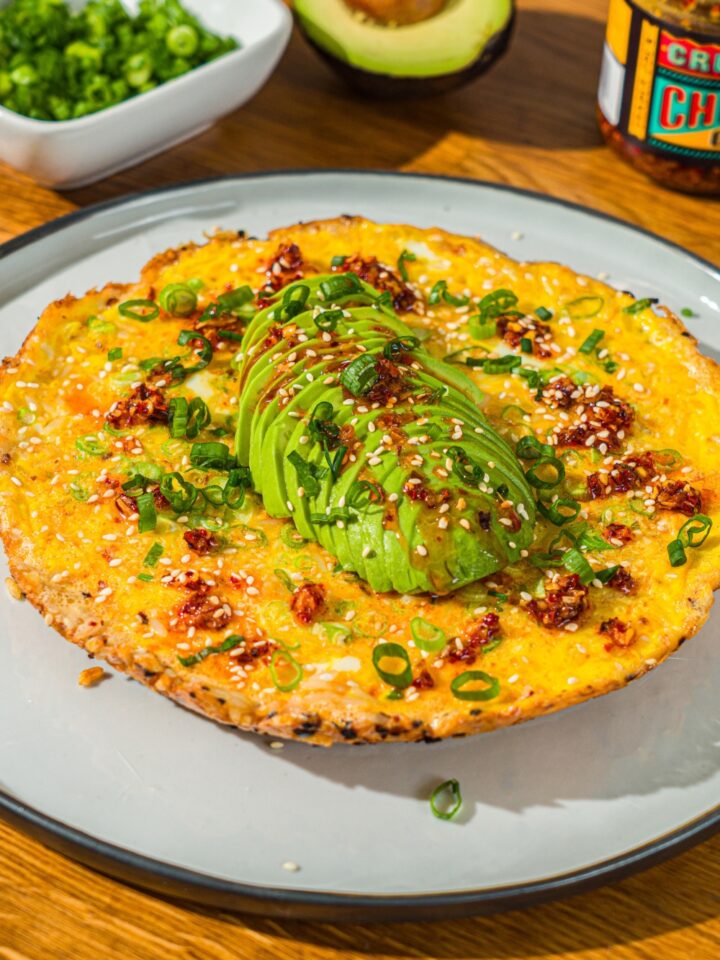 A white plate with a crispy rice omelette topped with sliced avocado, chili crunch oil, and sliced green onion. The plate is on a wooden counter with small bowls of garnishes and an avocado.