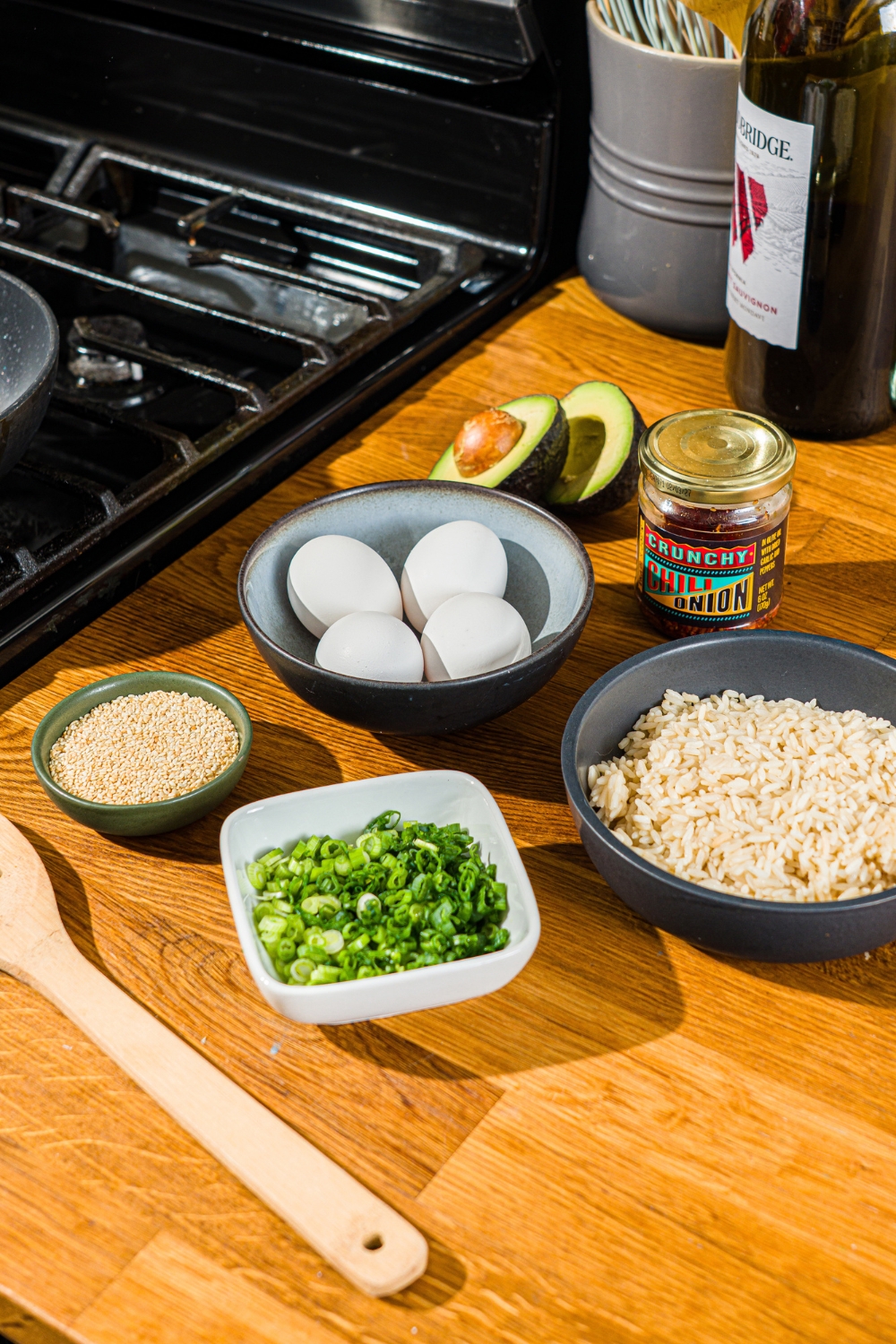 A wooden counter with several bowls containing ingredients to make crispy rice omelette including rice, egg, sesame seeds, avocado, and chili crunch oil