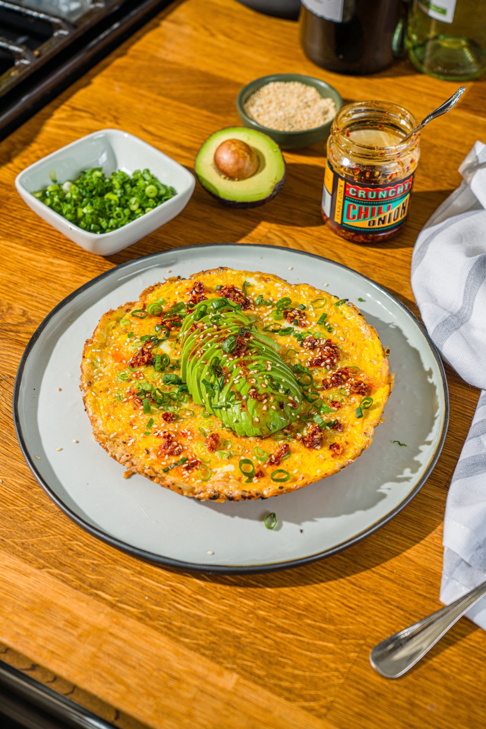 A white plate with a crispy rice omelette topped with sliced avocado, chili crunch oil, and sliced green onion. The plate is on a wooden counter with small bowls of garnishes and an avocado.