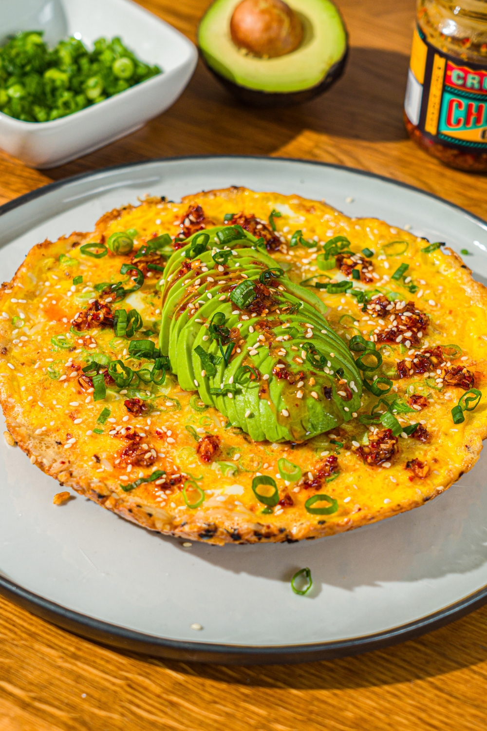 A white plate with a crispy rice omelette topped with sliced avocado, chili crunch oil, and sliced green onion. The plate is on a wooden counter with small bowls of garnishes and an avocado.