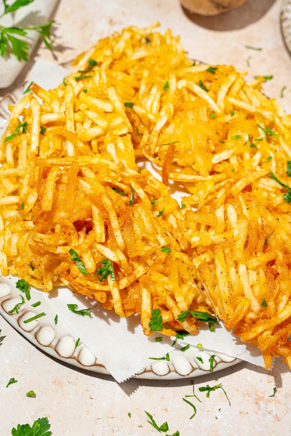 A plate lined with parchment paper with crispy parmesan hashbrowns garnished with fresh parsley. The plate is on a tan counter.