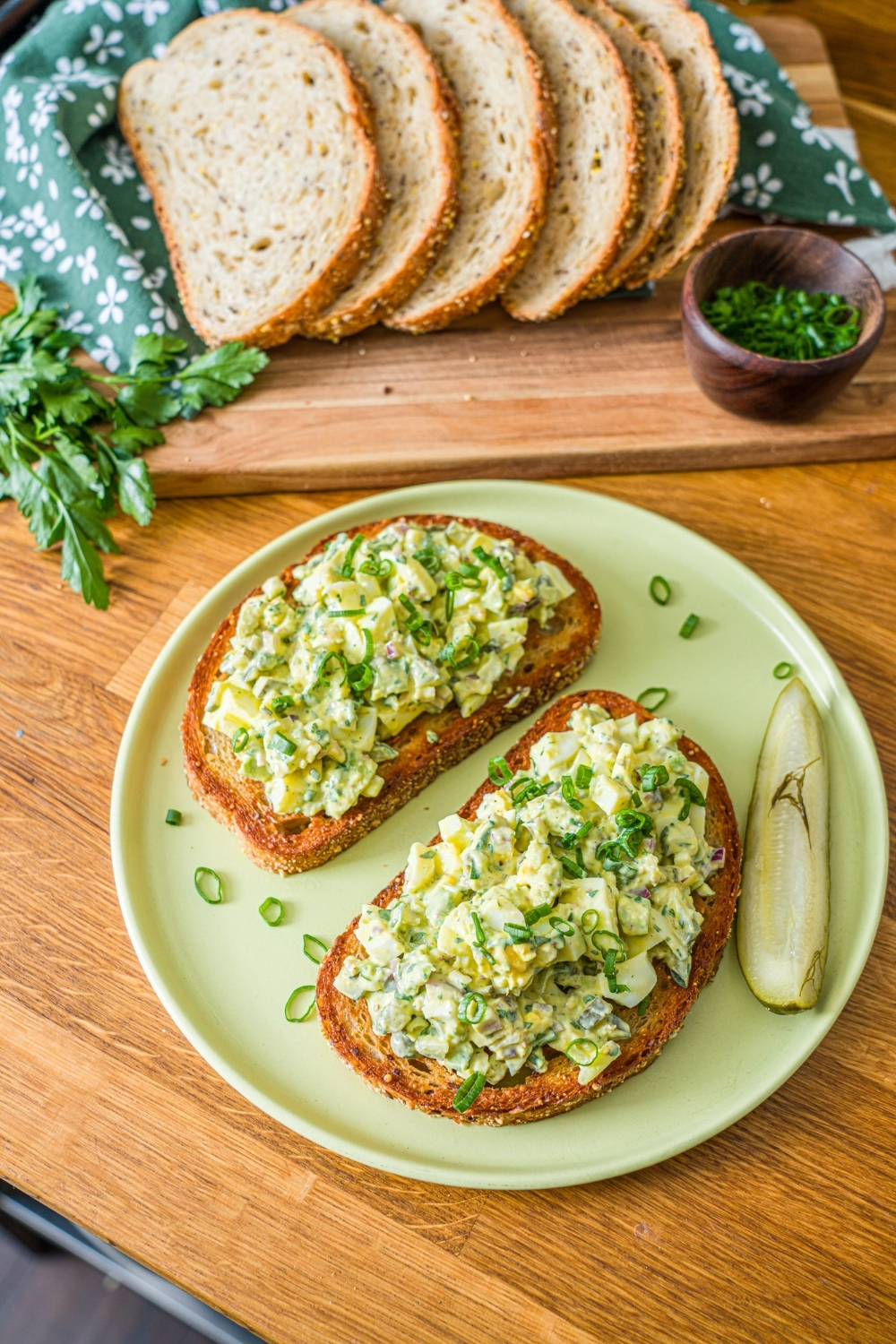 A green plate with two slices of toasted bread topped with pickle egg salad garnished with sliced green onion and served with a pickle spear. The plate is on a wooden counter with a wooden board of sliced bread with garnishes.