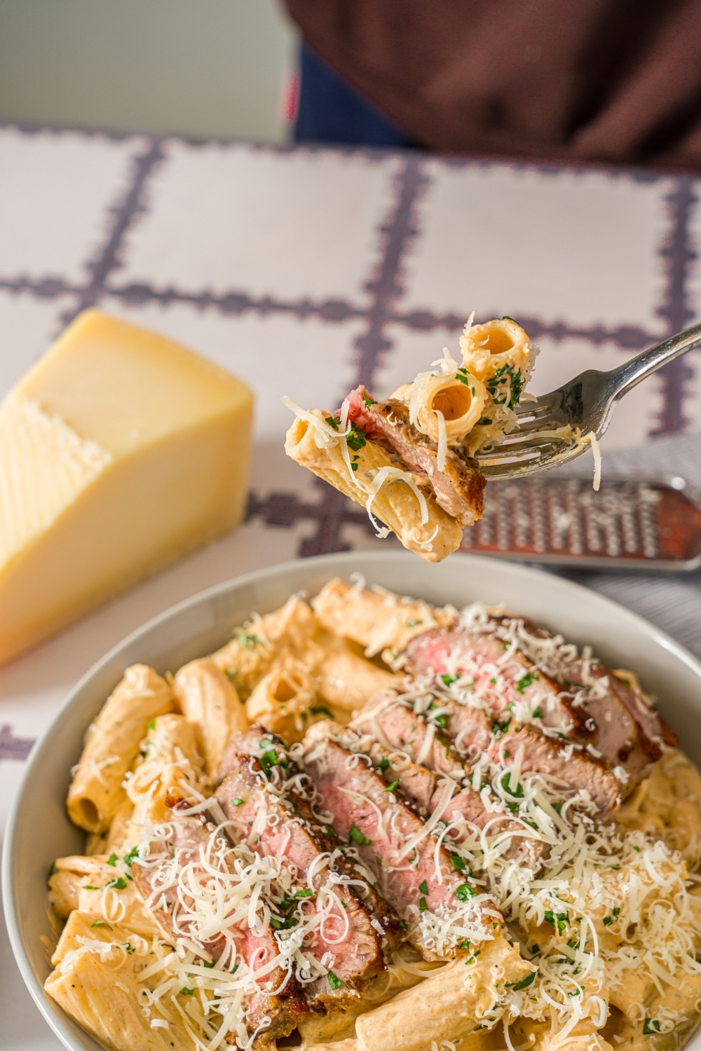 A fork with a bite of creamy cajun steak pasta garnished with fresh parsley and grated cheese. The fork is over a bowl of pasta. The bowl is on a tiled counter with a small bowl of parsley and block of parmesan cheese.