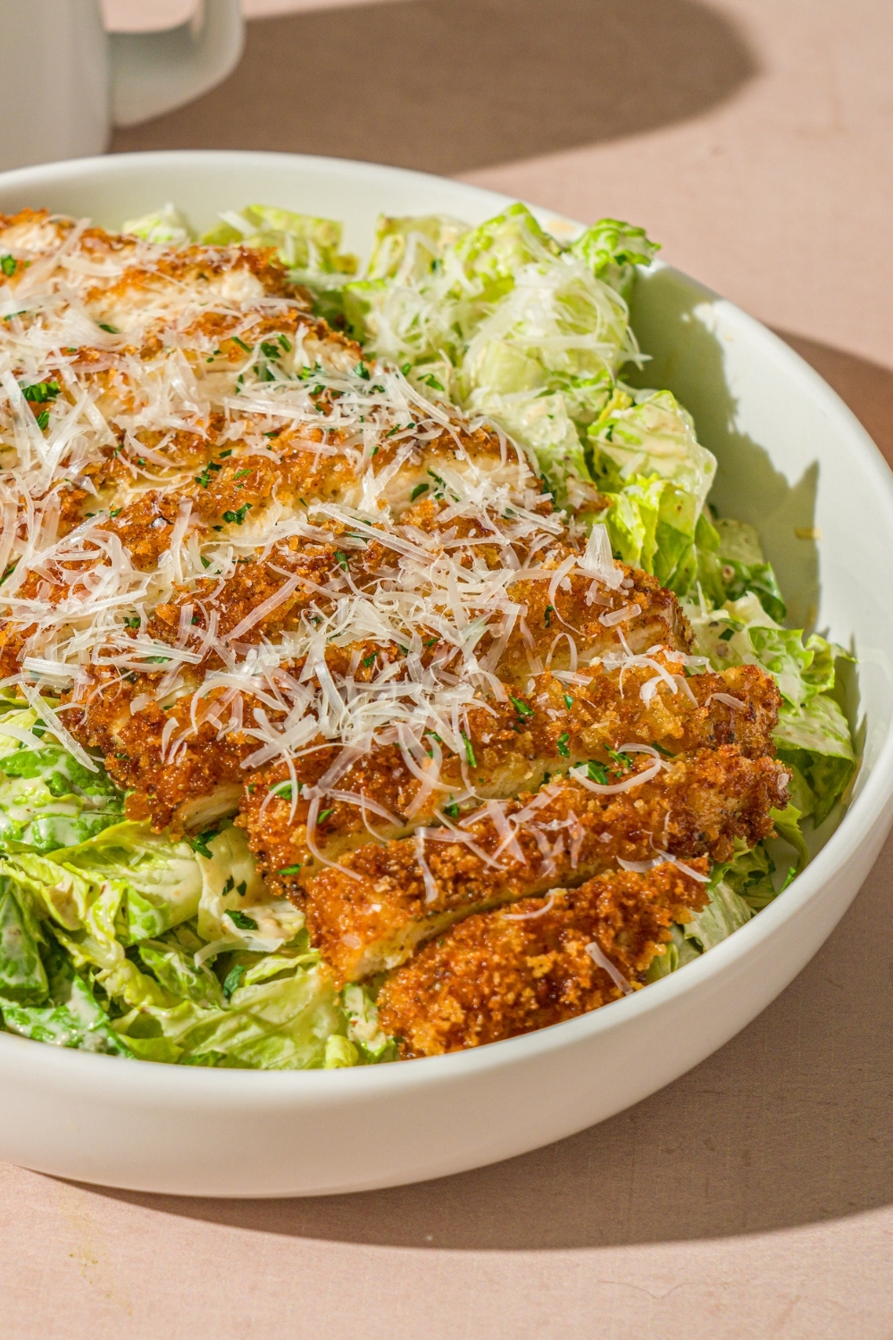 A white bowl with crispy chicken caesar salad garnished with fresh parsley and shredded parmesan. The bowl is on a tan counter.
