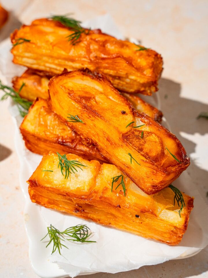 A white platter lined with parchment paper with several pieces of potato pave garnished with fresh thyme. The pieces are stacked on top of each other. The plate is on a tan counter.