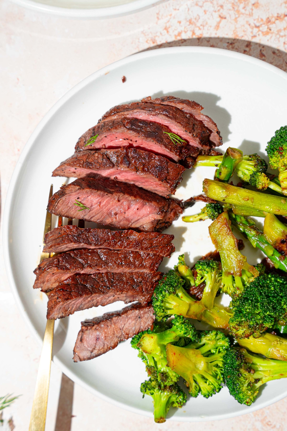 A white plate with sliced marinated skirt steak served with broccoli. There is a fork on the plate. The plate is on a tan counter.