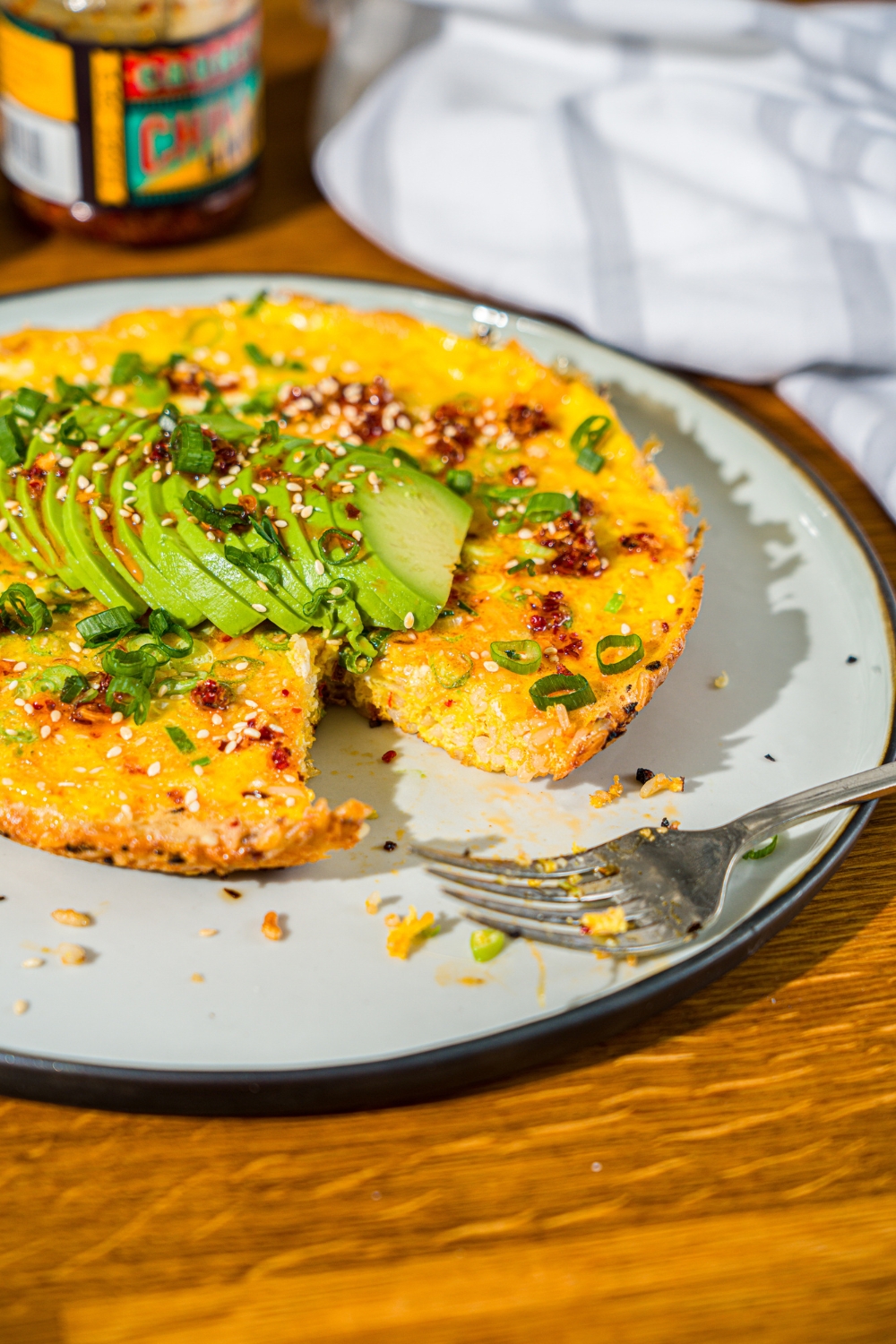 A white plate with a crispy rice omelette topped with sliced avocado, chili crunch oil, and sliced green onion. A bite is taken from the omelette. There is a fork on the plate. The plate is on a wooden counter with small bowls of garnishes and an avocado.
