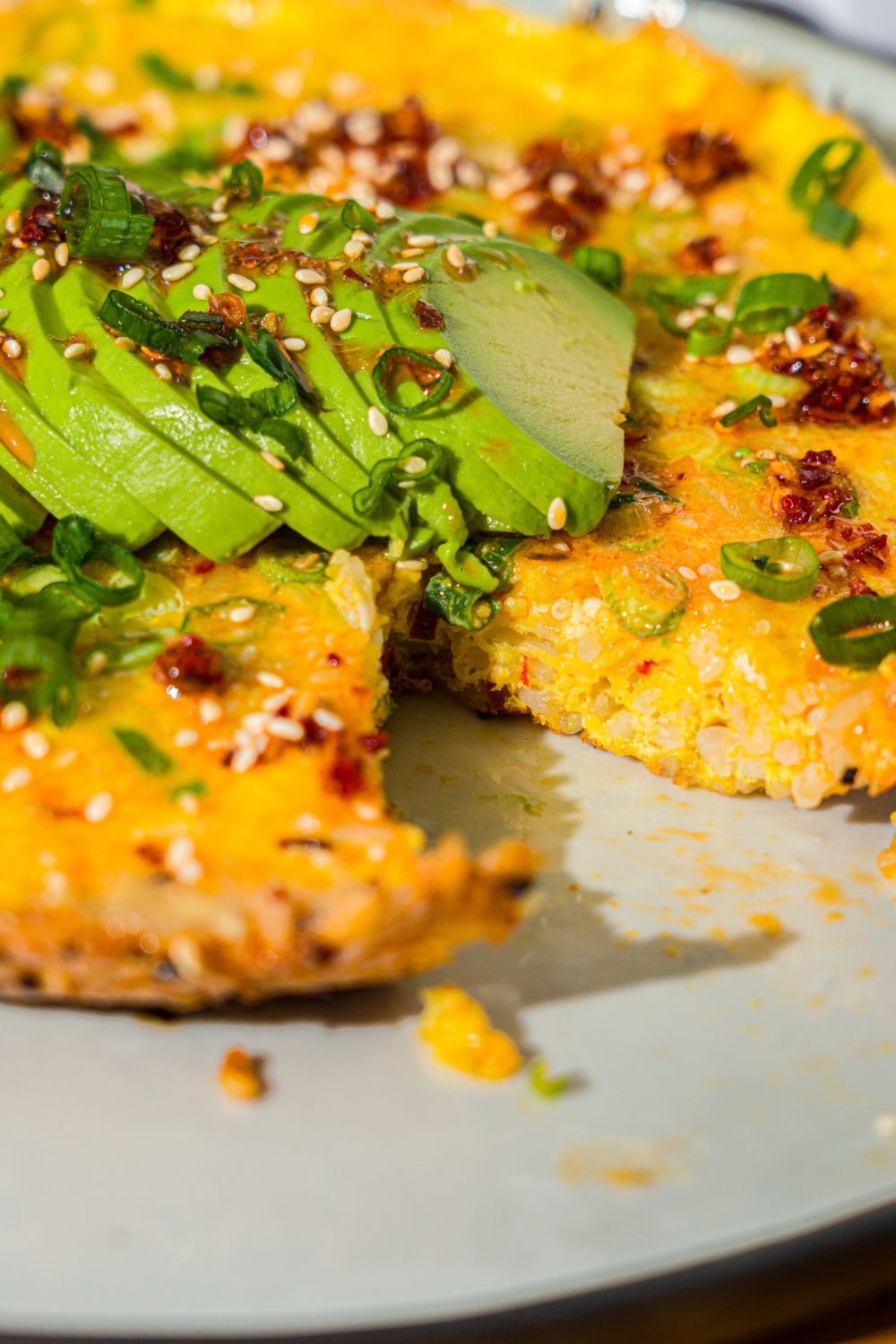 A white plate with a crispy rice omelette topped with sliced avocado, chili crunch oil, and sliced green onion. A bite is taken from the omelette.