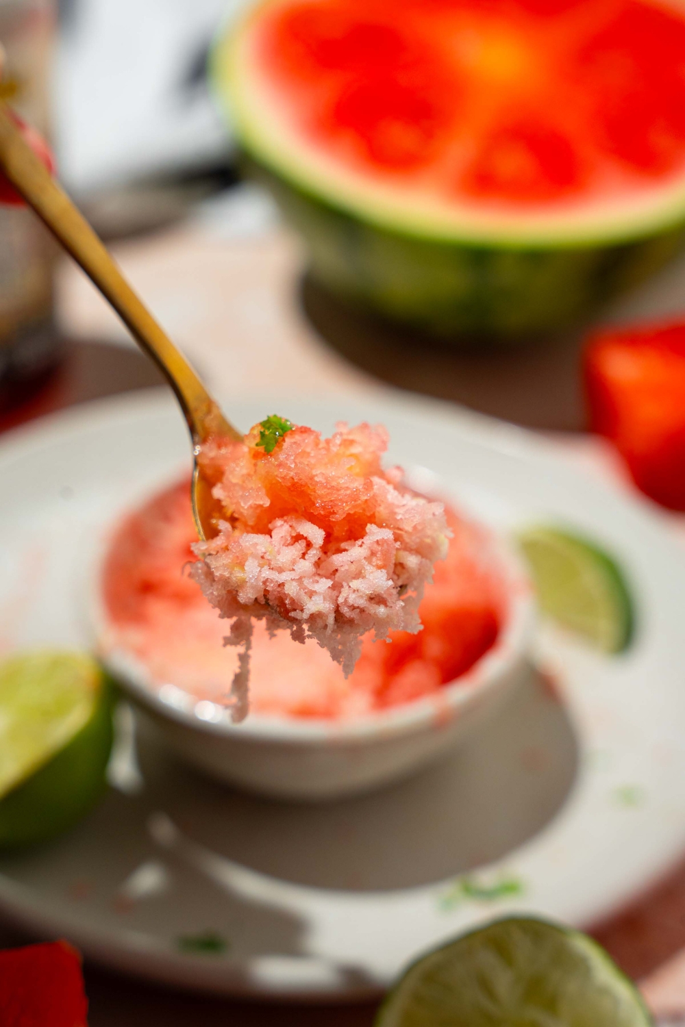 A close up of a spoon with a bite of watermelon shaved ice. There is a bowl of shaved ice on a white plate blurred in the background.
