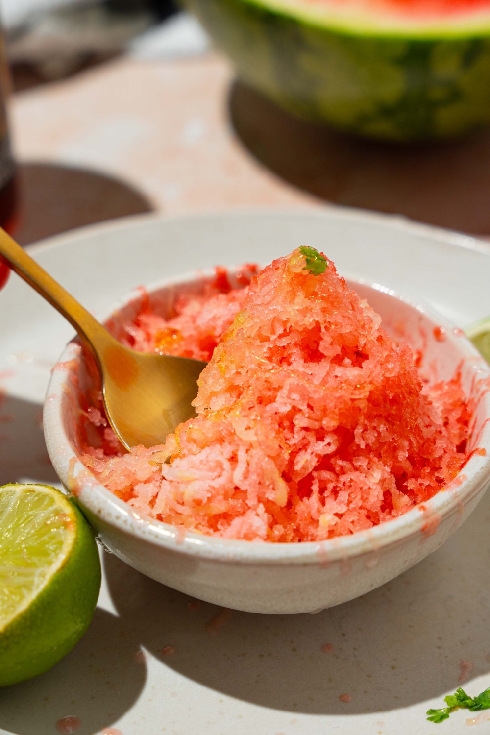A small white bowl with watermelon shaved ice drizzled with honey. There is a spoon in the bowl. The bowl is on a white plate with lime.