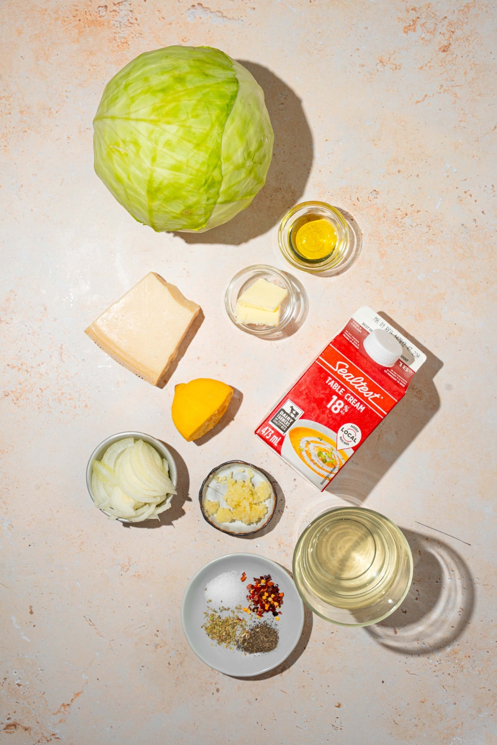 A tan counter with several bowls in various sizes containing ingredients to make garlic parmesan melted cabbage including cabbage, heavy cream, butter, parmesan cheese, garlic, chicken broth, onion, olive oil, and seasonings.