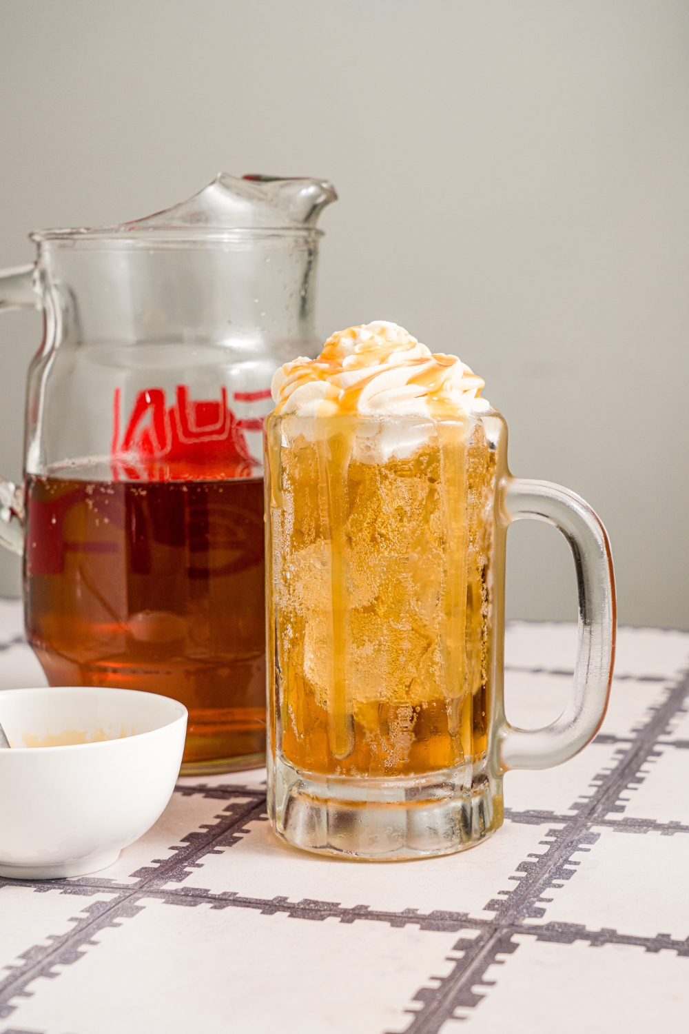 A large beer glass with butterbeer topped with butterscotch whipped cream. Butterscotch syrup is being drizzled over the glass. The glass is on a tiled counter with a small bowl of butterscotch syrup.