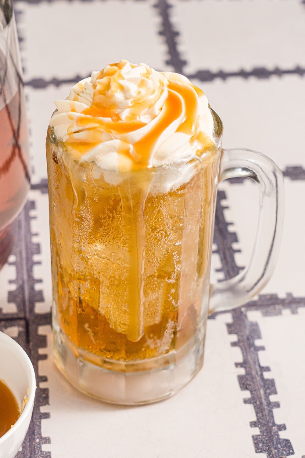 A large beer glass with butterbeer topped with butterscotch whipped cream. Butterscotch syrup is being drizzled over the glass. The glass is on a tiled counter with a small bowl of butterscotch syrup.