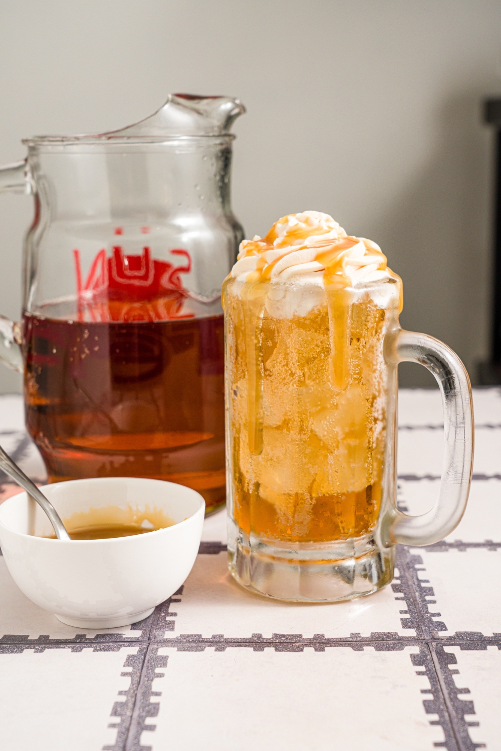 A large beer glass with butterbeer topped with butterscotch whipped cream. Butterscotch syrup is being drizzled over the glass. The glass is on a tiled counter with a small bowl of butterscotch syrup.