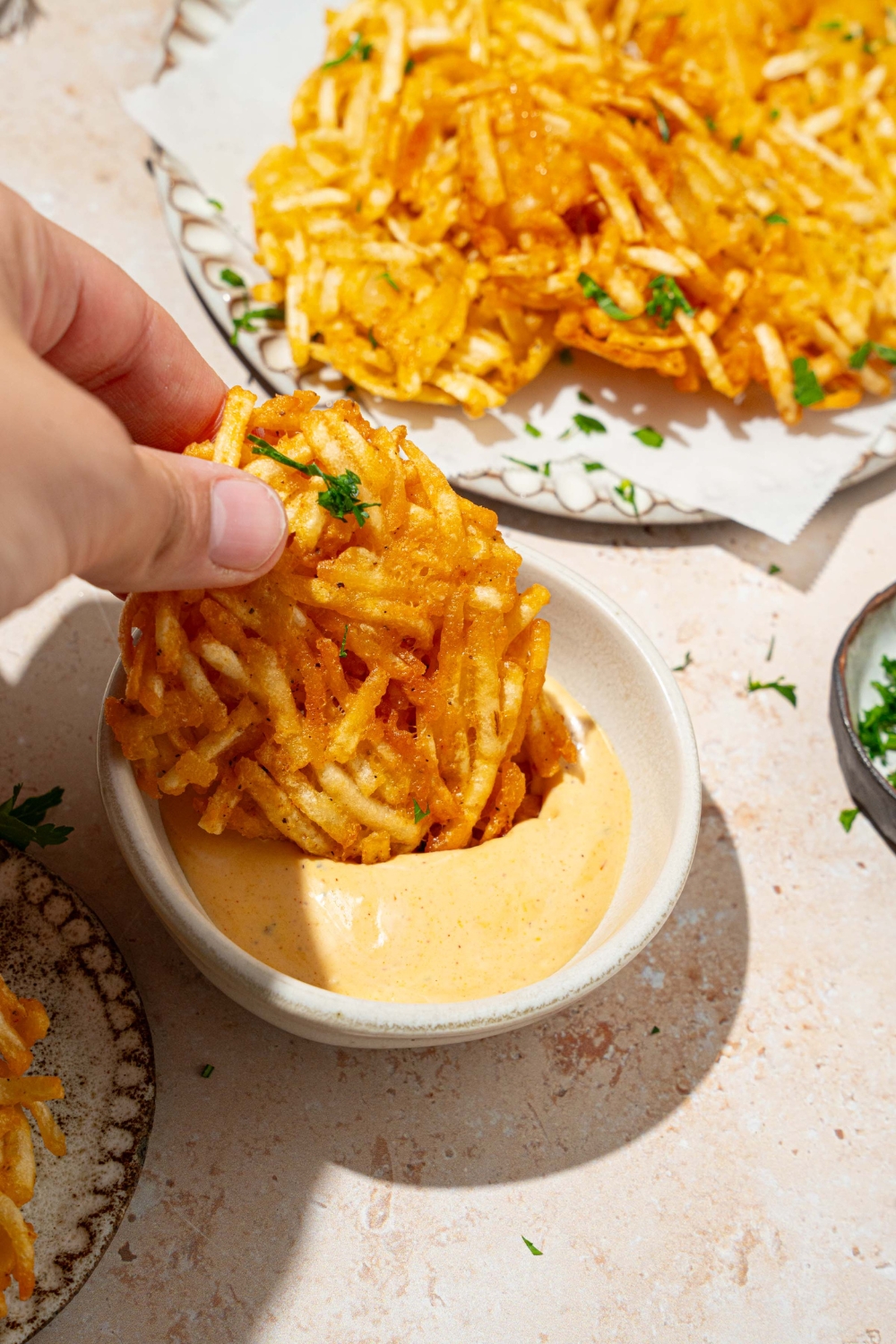 A hand dipping a crispy parmesan hashbrown into a bowl of dipping sauce. The bowl is on a tan counter with a plate of hashbrowns.