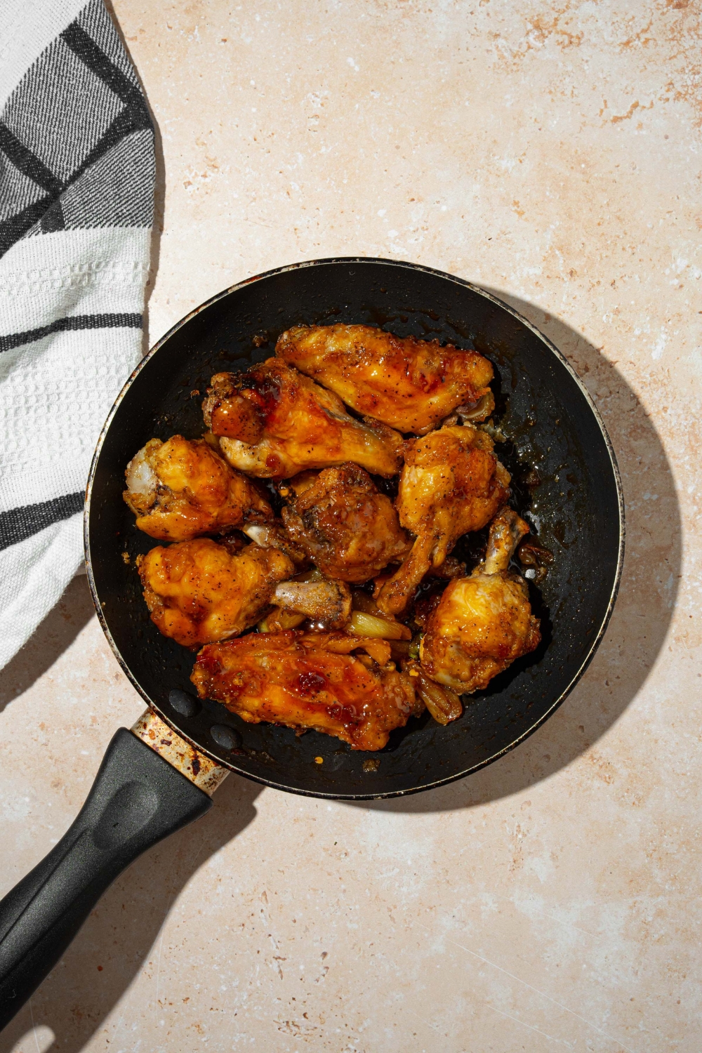 A skillet with baked chicken wings tossed in Asian zing sauce. The skillet is on a tan counter with a gray cloth napkin.