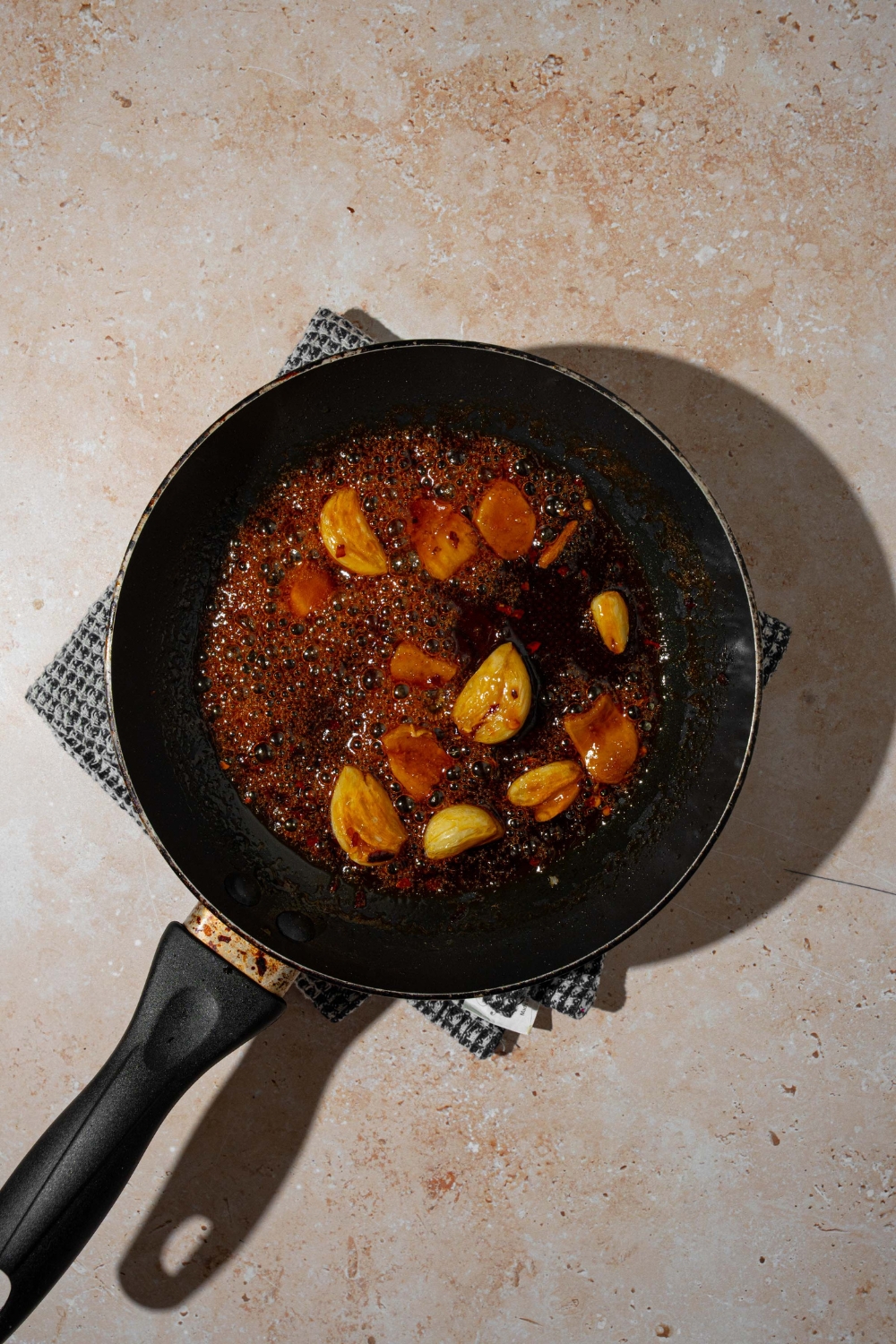 A skillet with smashed garlic cooking in Asian zing sauce mixture. The skillet is on a tan counter with a gray cloth napkin.