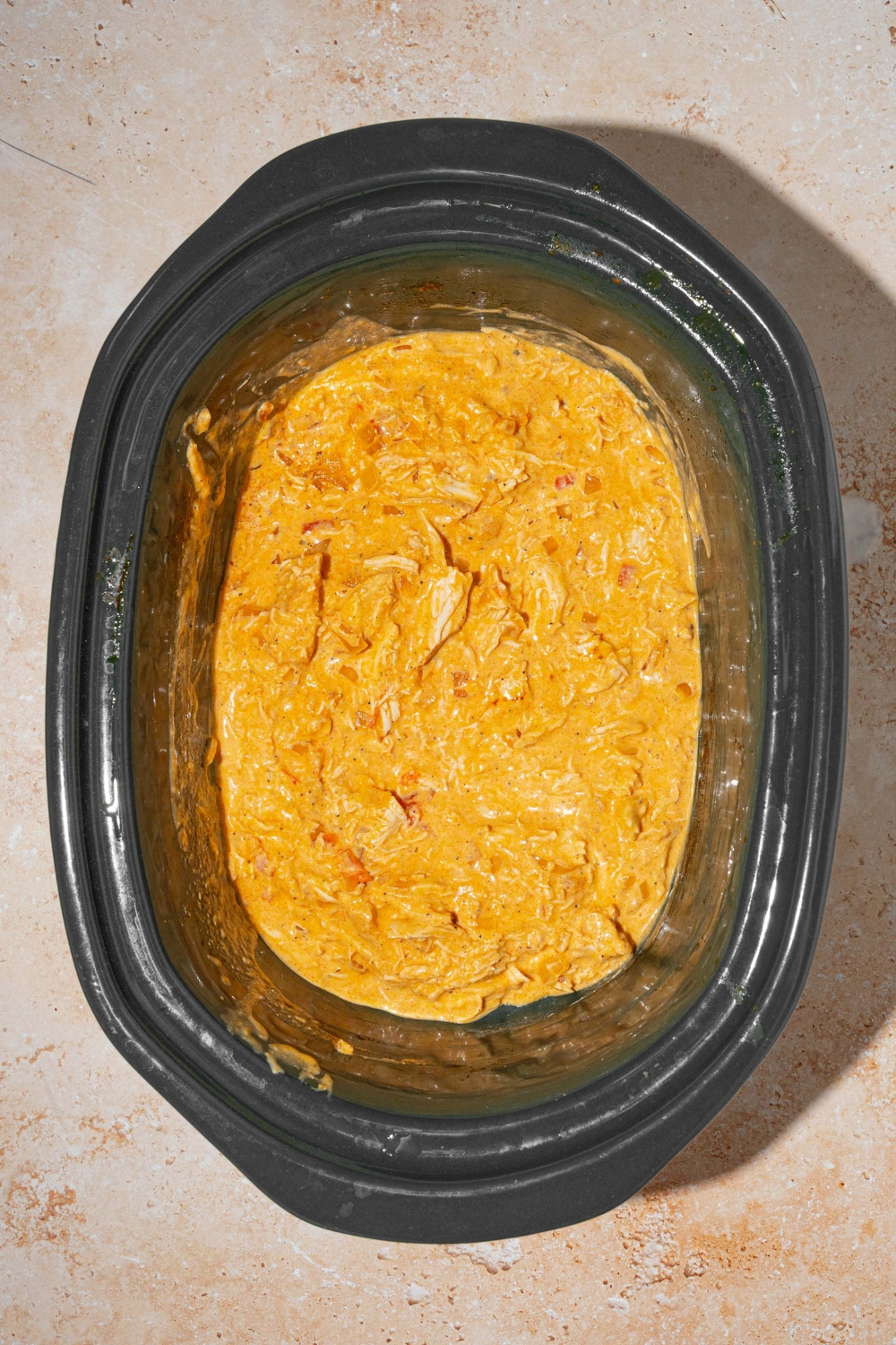 A slow cooker with shredded chicken tossed in a Buffalo cheese sauce. The slow cooker is on a tan counter.