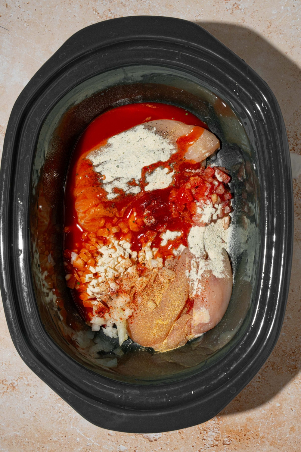 A slow cooker with uncooked chicken and additional ingredients to make Buffalo chicken including Buffalo sauce and seasonings.
