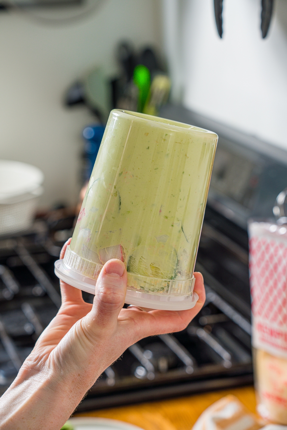 A hand shaking a sealed deli container with ingredients to make California roll cucumber salad including sliced cucumber and imitation crab with an avocado cream cheese dressing.