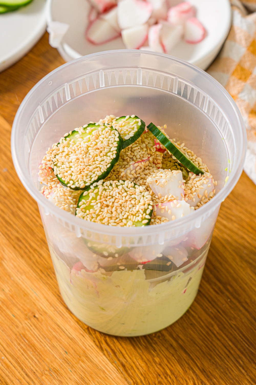A deli container with ingredients to make California roll cucumber salad including sliced cucumber, imitation crab, and sesame seeds with an avocado cream cheese dressing. The container is on a wooden counter.