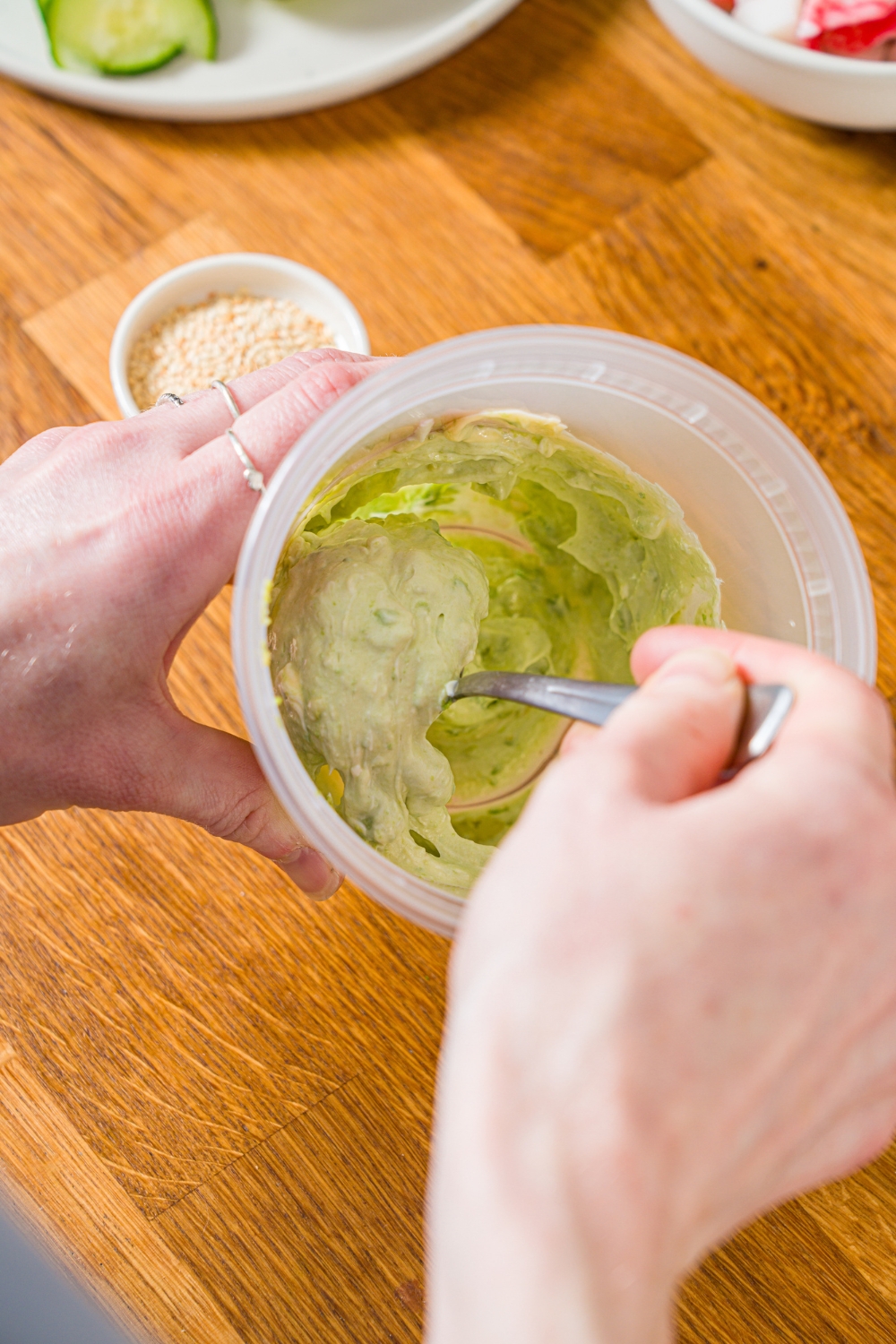 A spoon mixing an avocado cream cheese mixture in a deli container. The container is on a wooden counter.