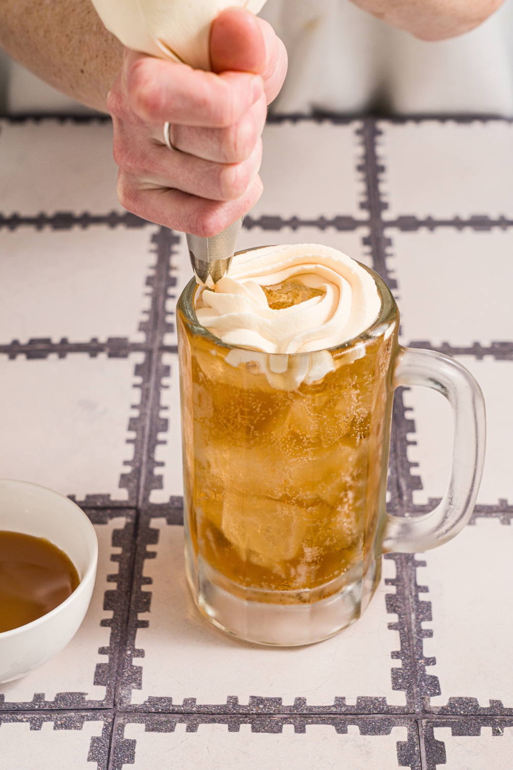 A large beer glass with butterbeer. A piping bag is piping butterscotch whipped cream over the butterbeer. The glass is on a tiled counter.