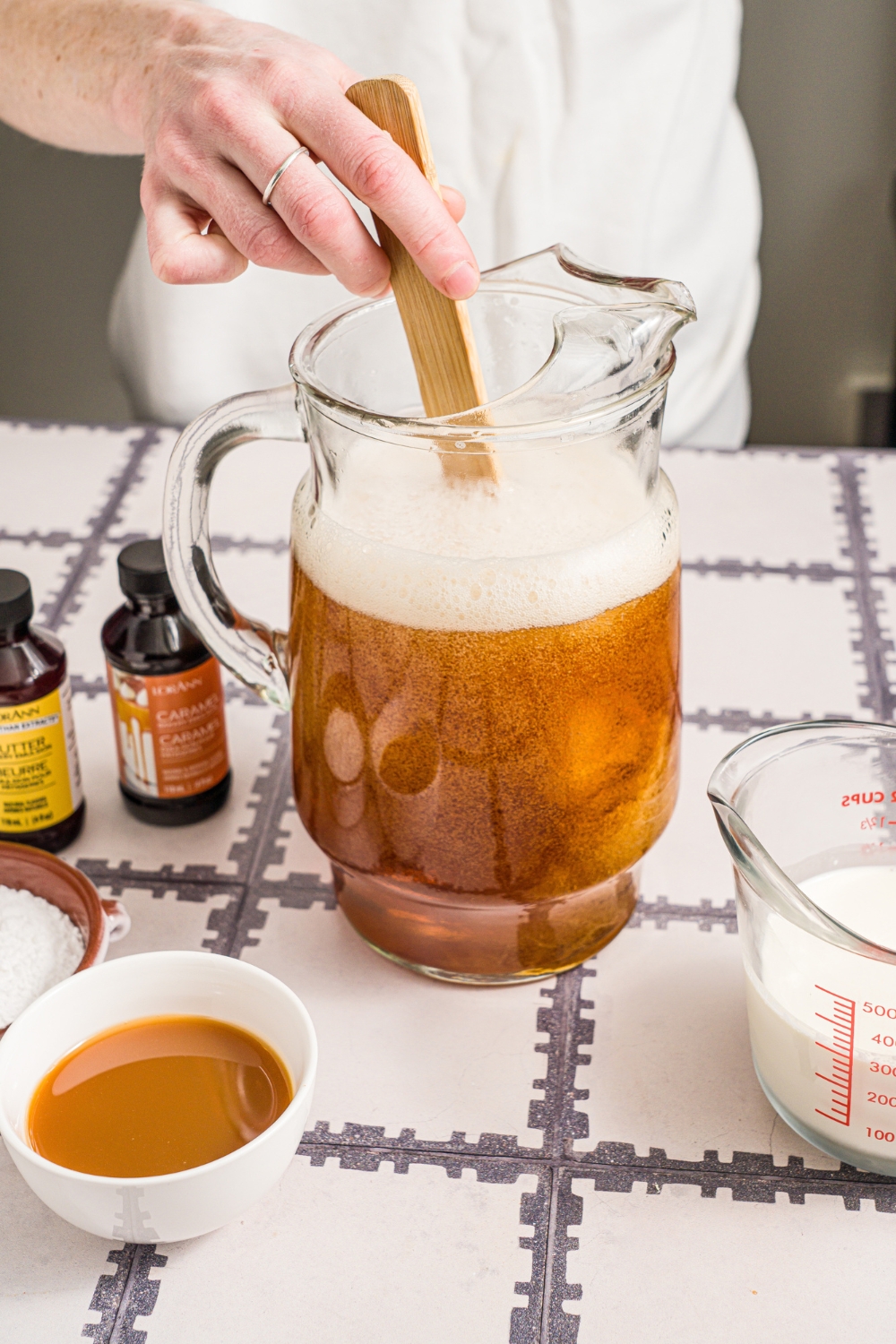 A large pitcher with cream soda, butter extract, and caramel extract. A spoon is stirring the mixture. The pitcher is on a tiled counter with ingredients.
